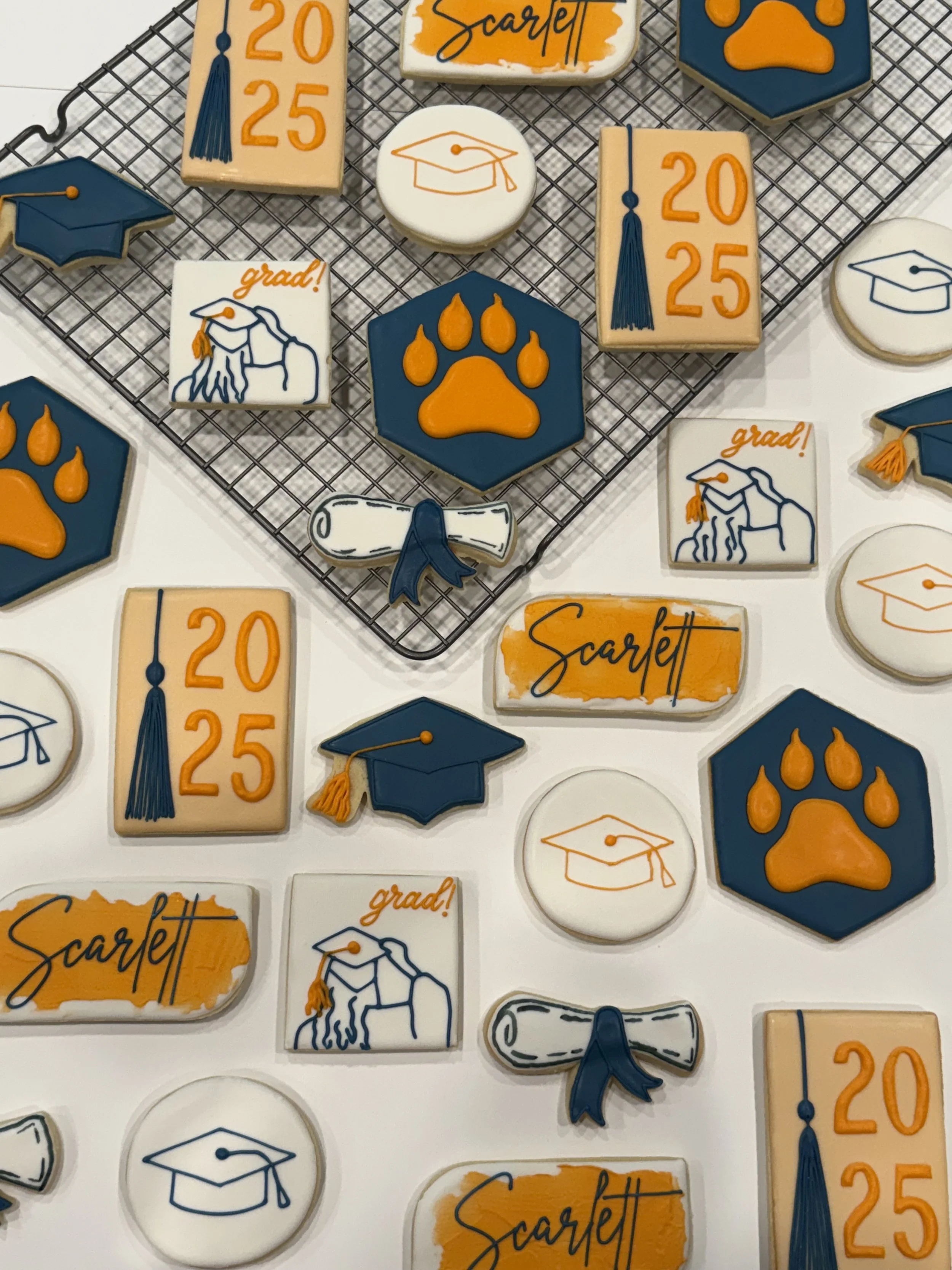 A collection of decorated graduation cookies with various designs, including cap and diploma motifs, paw prints, and the name 'Scarlett' written in orange and blue.