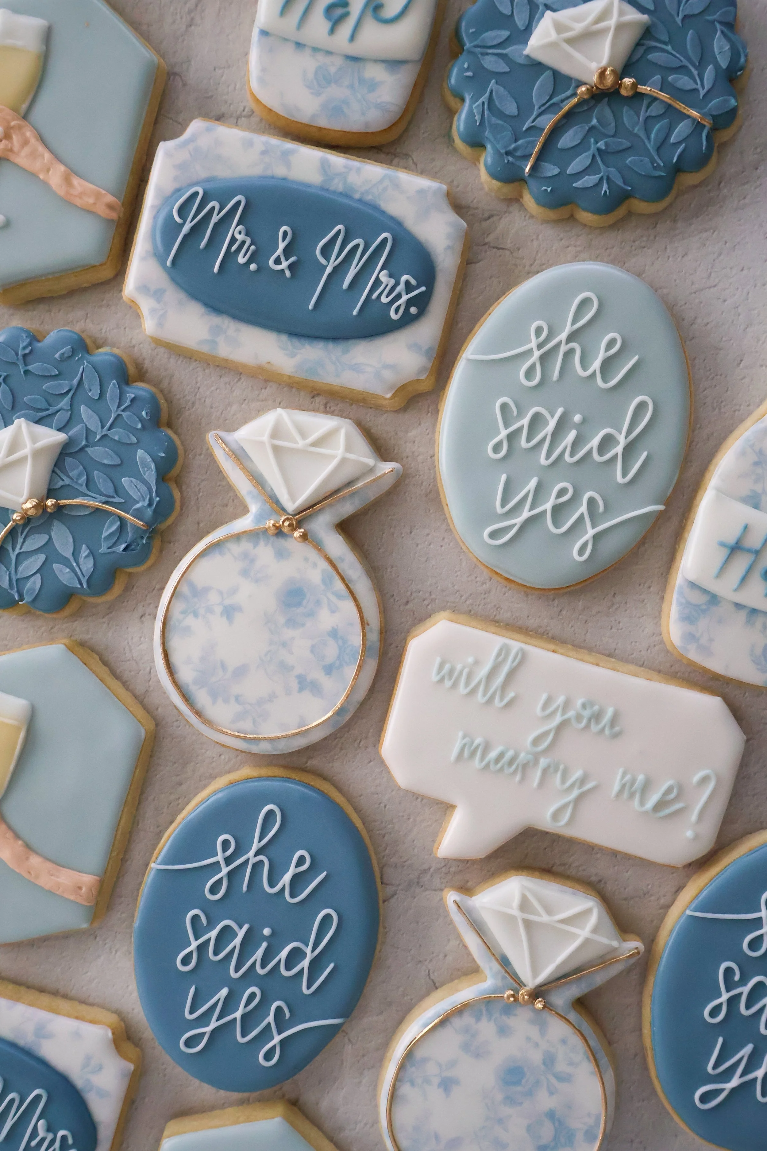 Collection of blue, white, and cream decorated cookies with wedding-themed messages and symbols, including cookies with words "Mr. & Mrs." and "she said yes," along with diamond ring designs and floral patterns.