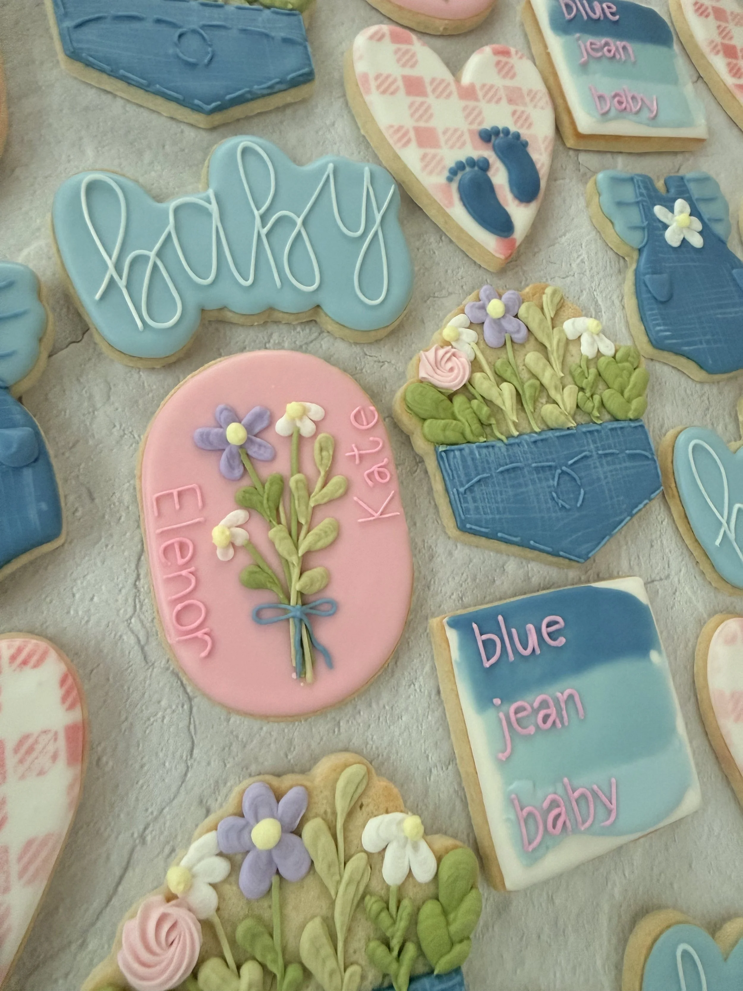 Decorated cookies with pastel colors and words like "baby" and "blue jean"; some cookies are shaped like hearts and flowers, with detailed icing designs including flowers, footprints, and phrases.
