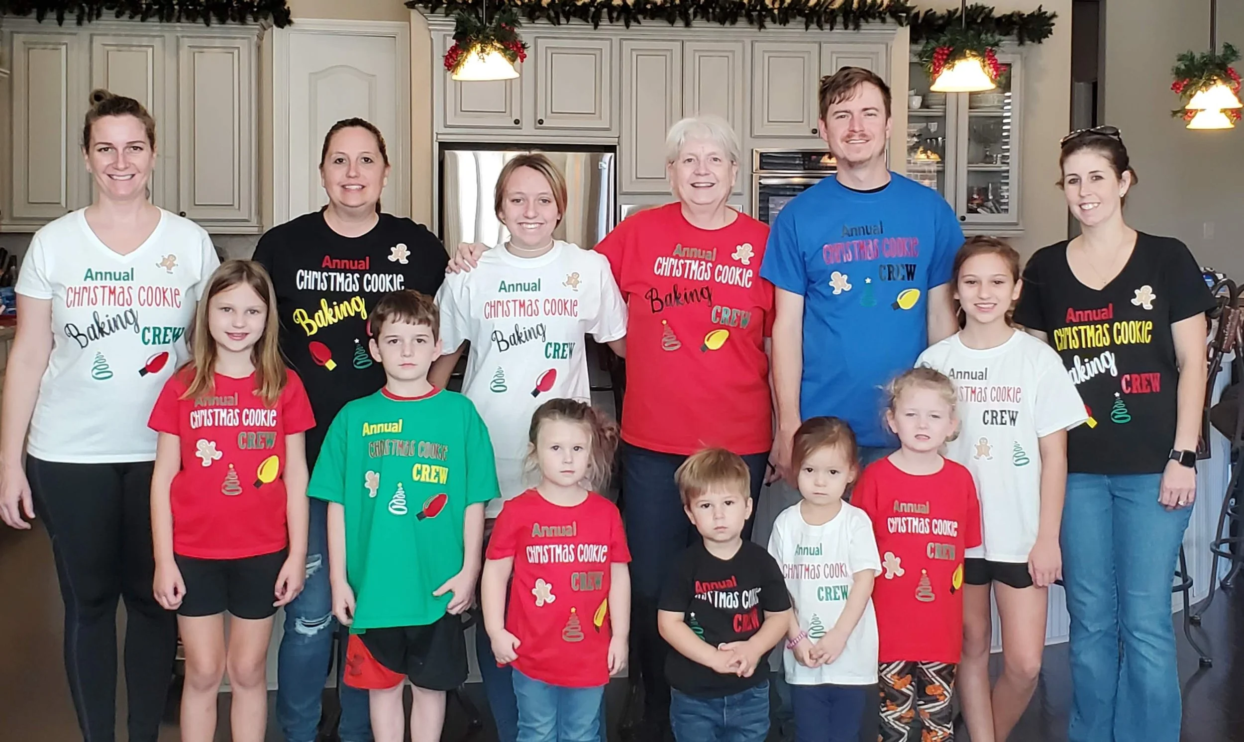 A group of people, including children and adults, standing together in a kitchen decorated for Christmas, wearing T-shirts with a Christmas cookie baking theme.
