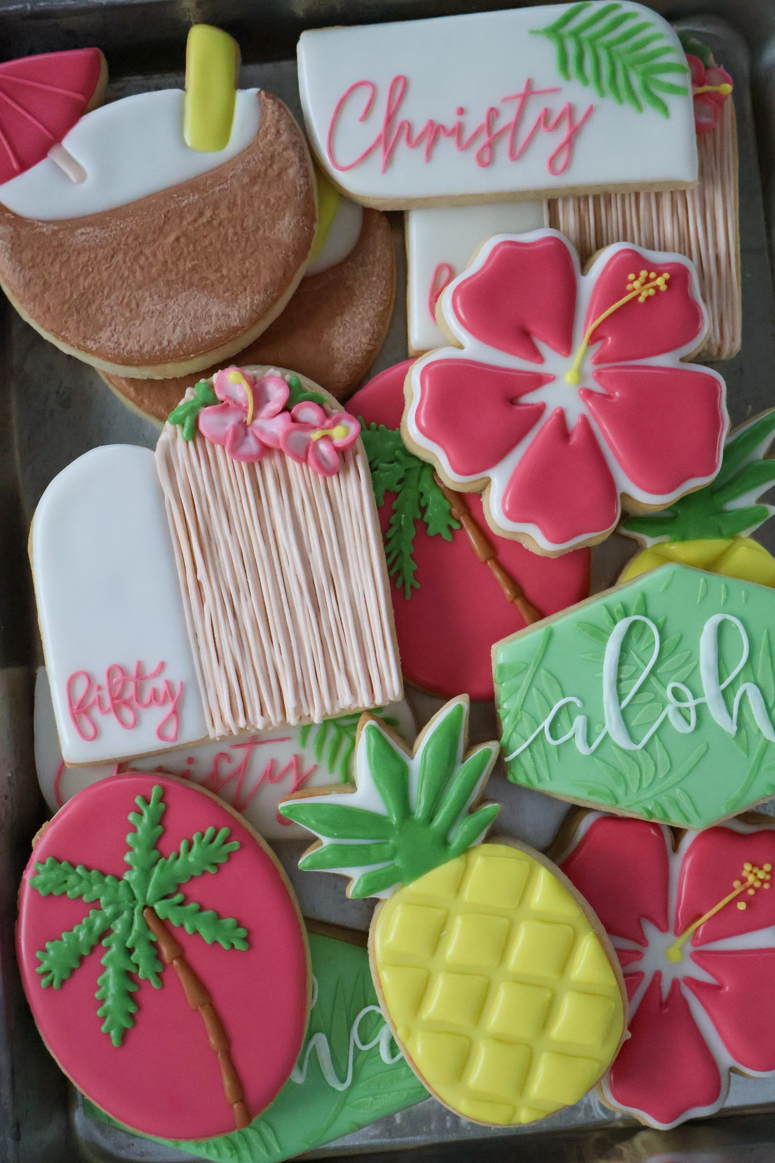 Colorful tropical-themed decorated cookies with flowers, pineapples, and trees, some with the word "Christy" written in pink icing.