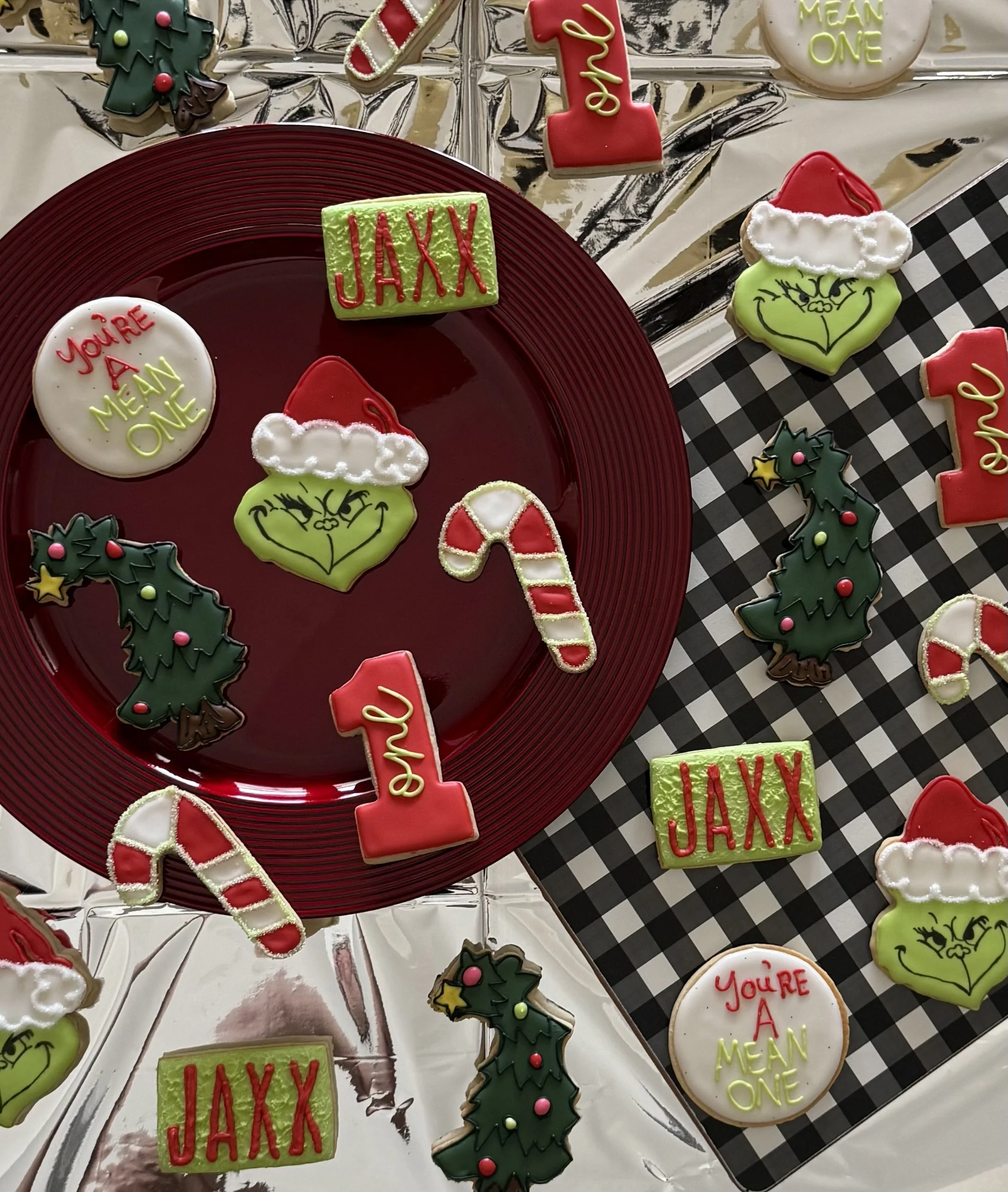 Arrangement of decorated Christmas cookies on a table, featuring holiday-themed shapes with red, green, and white icing, including holly leaves, candy canes, Santa hats, and cookies with messages like "You're a Christmas" and "Jaxx."