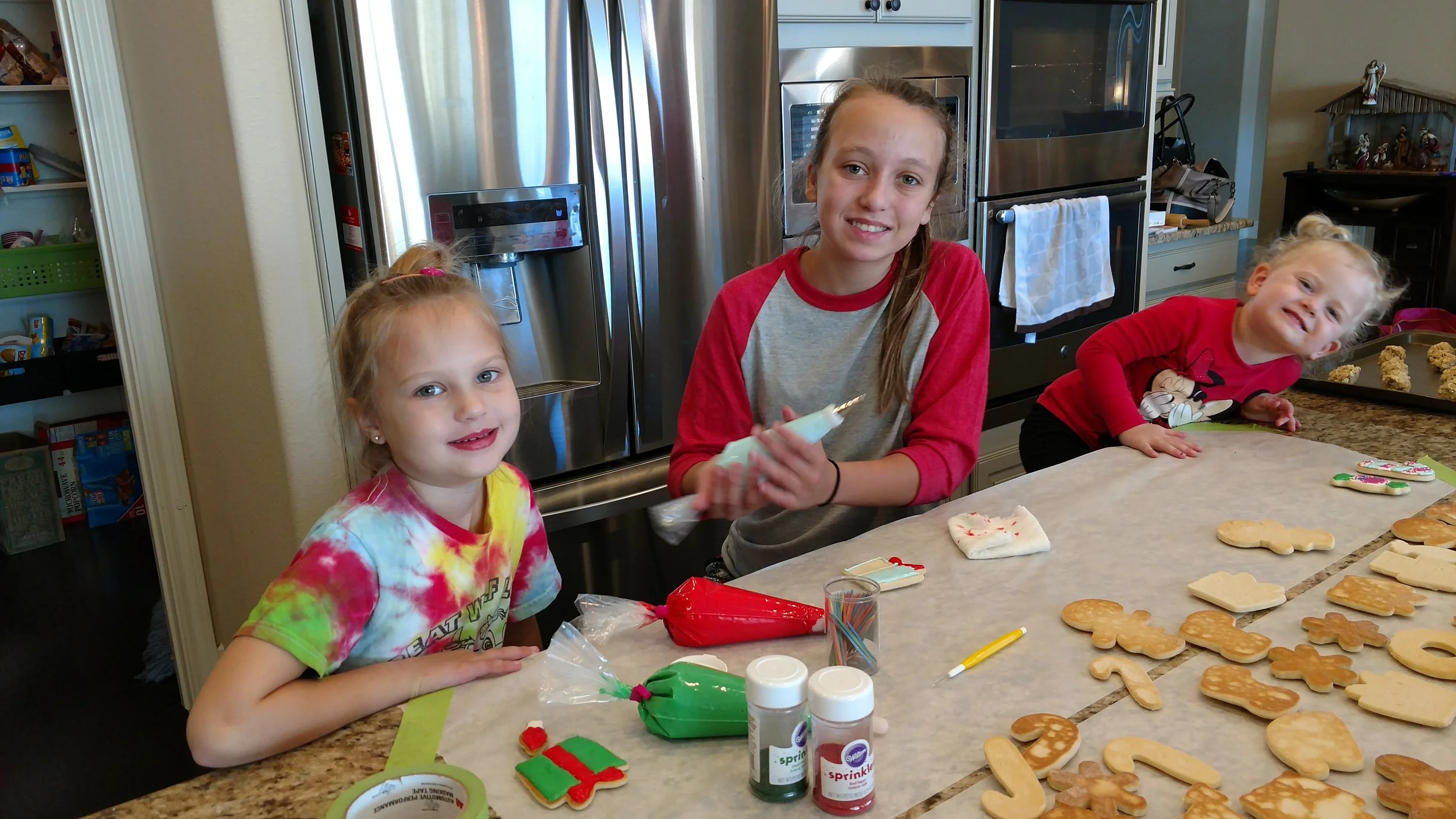 Three girls decorating Christmas cookies in a kitchen, with decorated cookies and baking supplies on the counter.