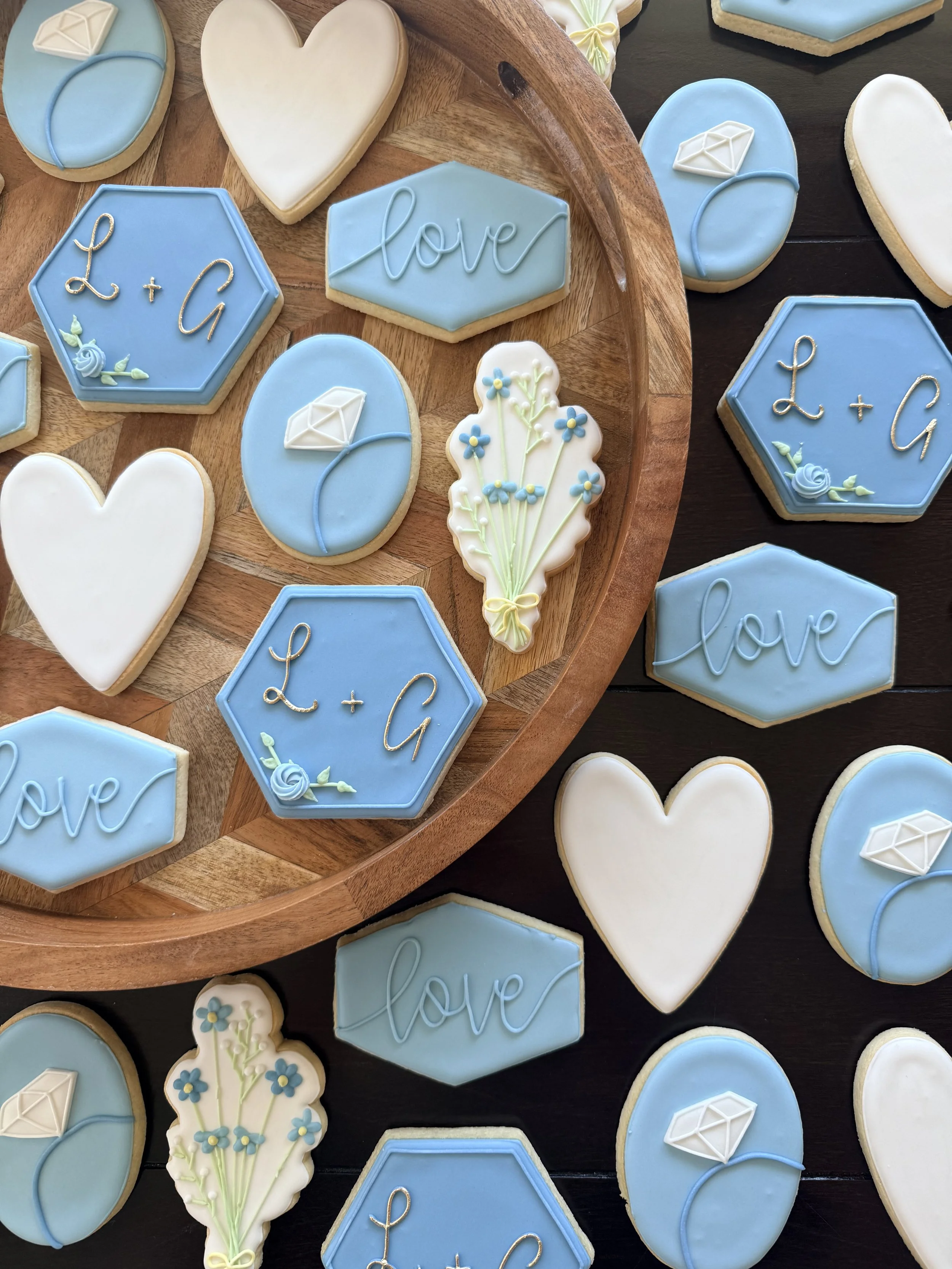 Decorated cookies with pastel blue and white icing, some featuring the word 'love' in cursive, hearts, diamonds, and floral designs, arranged on a wooden and dark surface.