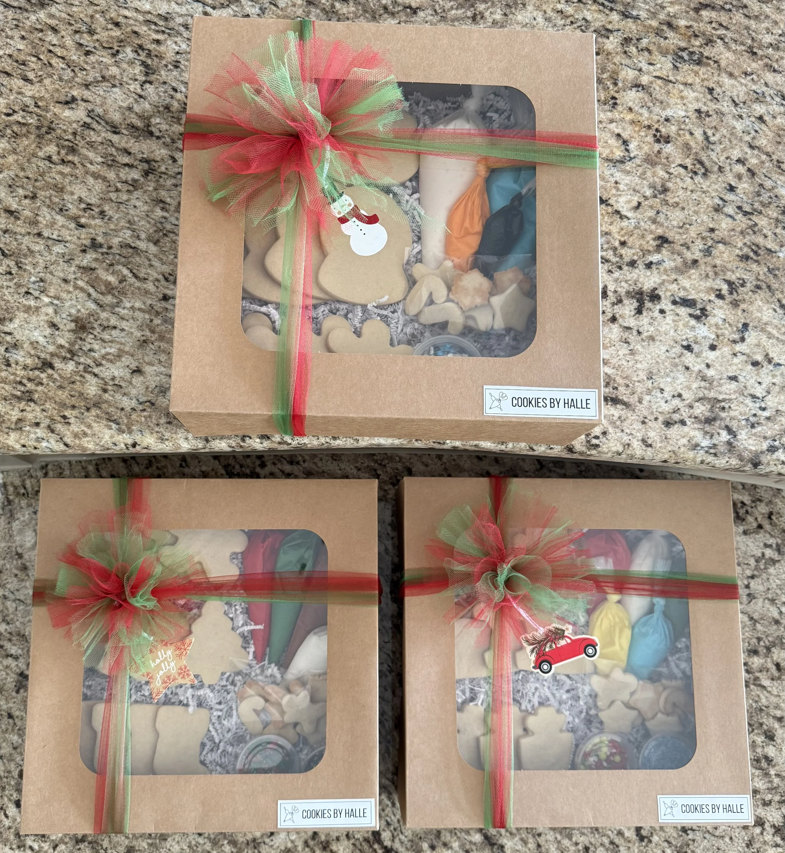 Boxes of holiday-themed cookies decorated with icing and sprinkles, each box wrapped with red and green ribbon and adorned with a small snowman and holiday-themed tags.