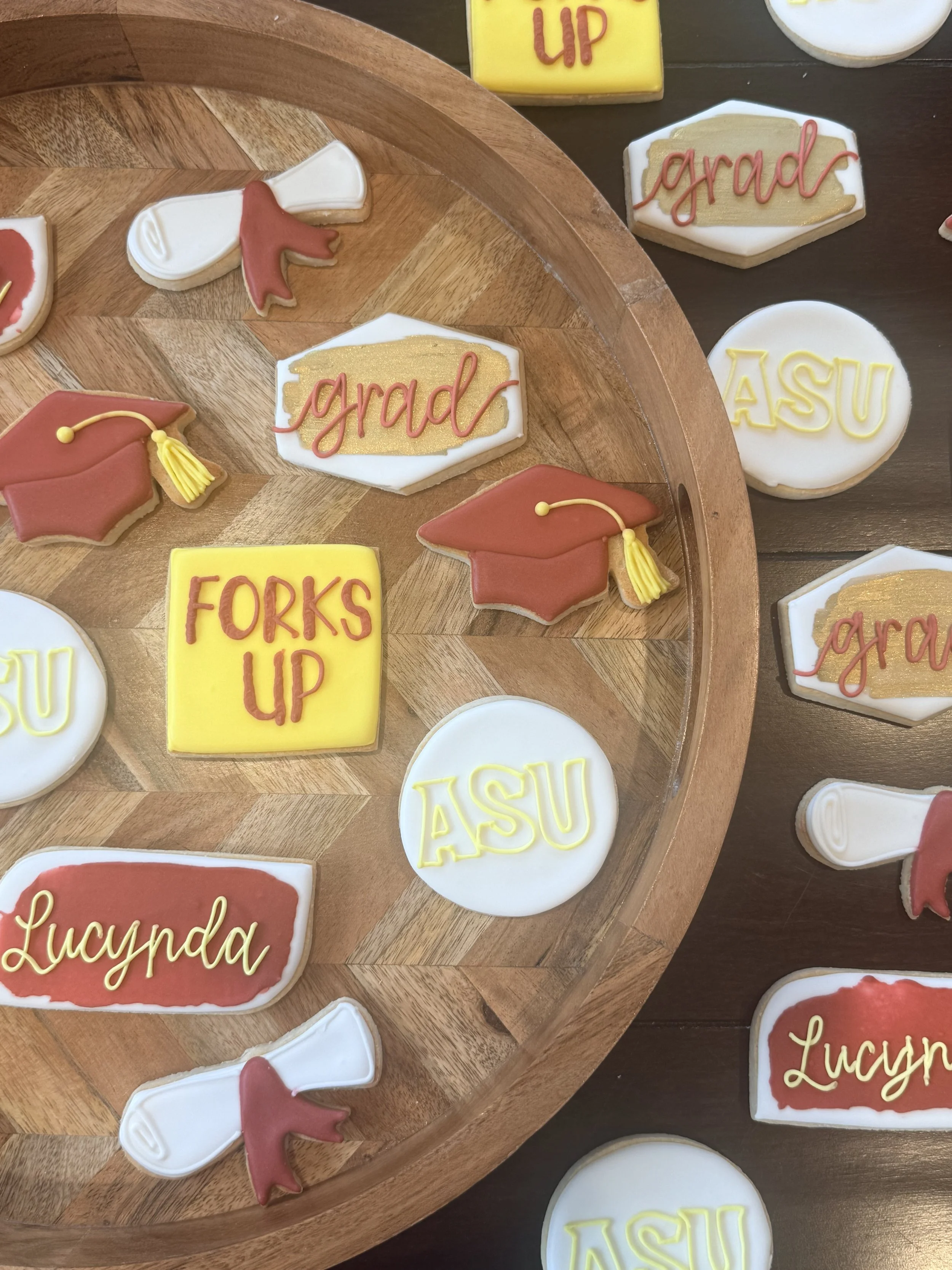 Cookies decorated with university campus names and graduation caps, including 'grad', 'FORKS UP', and 'ASU' written in colorful icing.
