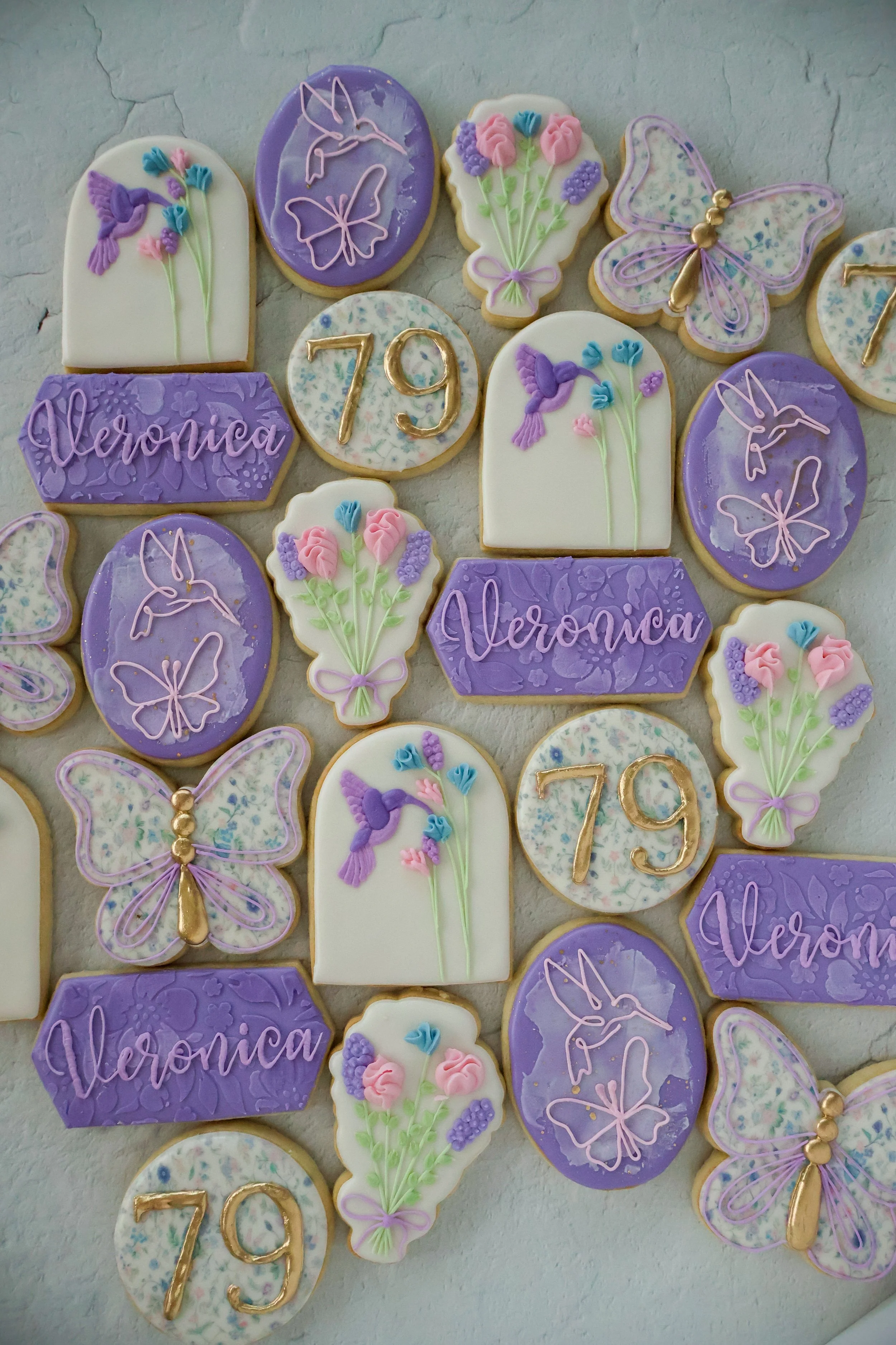 Assorted decorated cookies with purple, white, and pink designs. Some cookies have floral patterns, butterflies, and hummingbirds, with the words "Veronica" and the number "79" written on others.