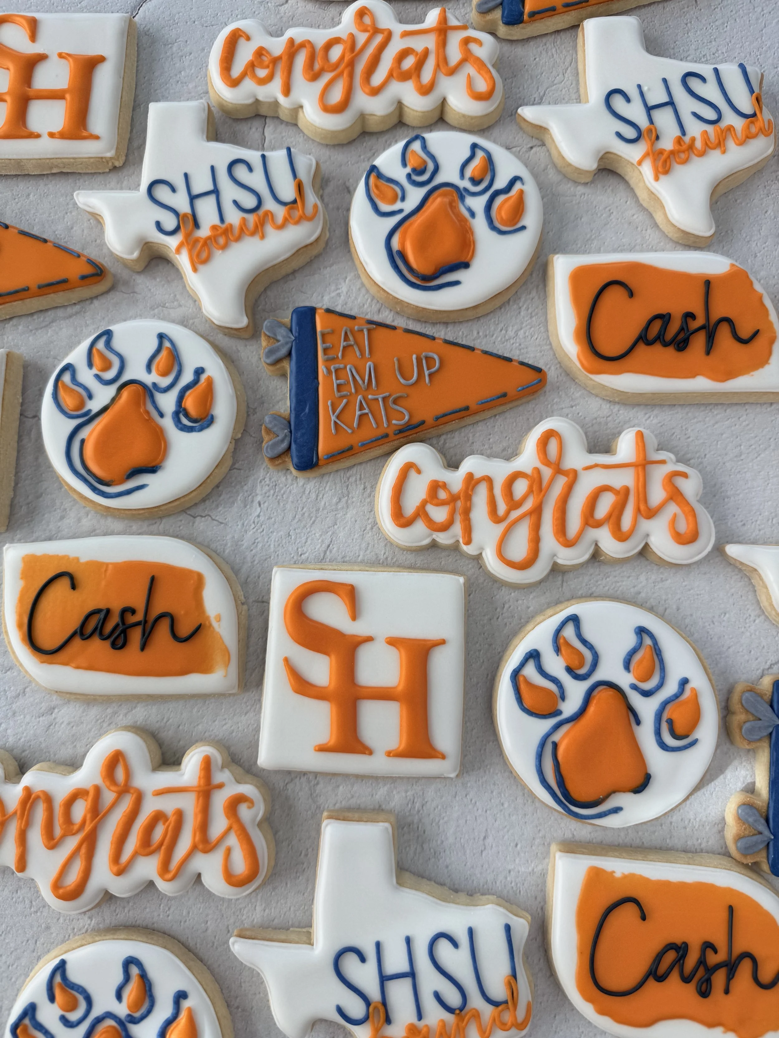 Decorated cookies with Texas shapes and Texas-themed messages like "Congrats" and "Cash" in orange, white, and blue icing.
