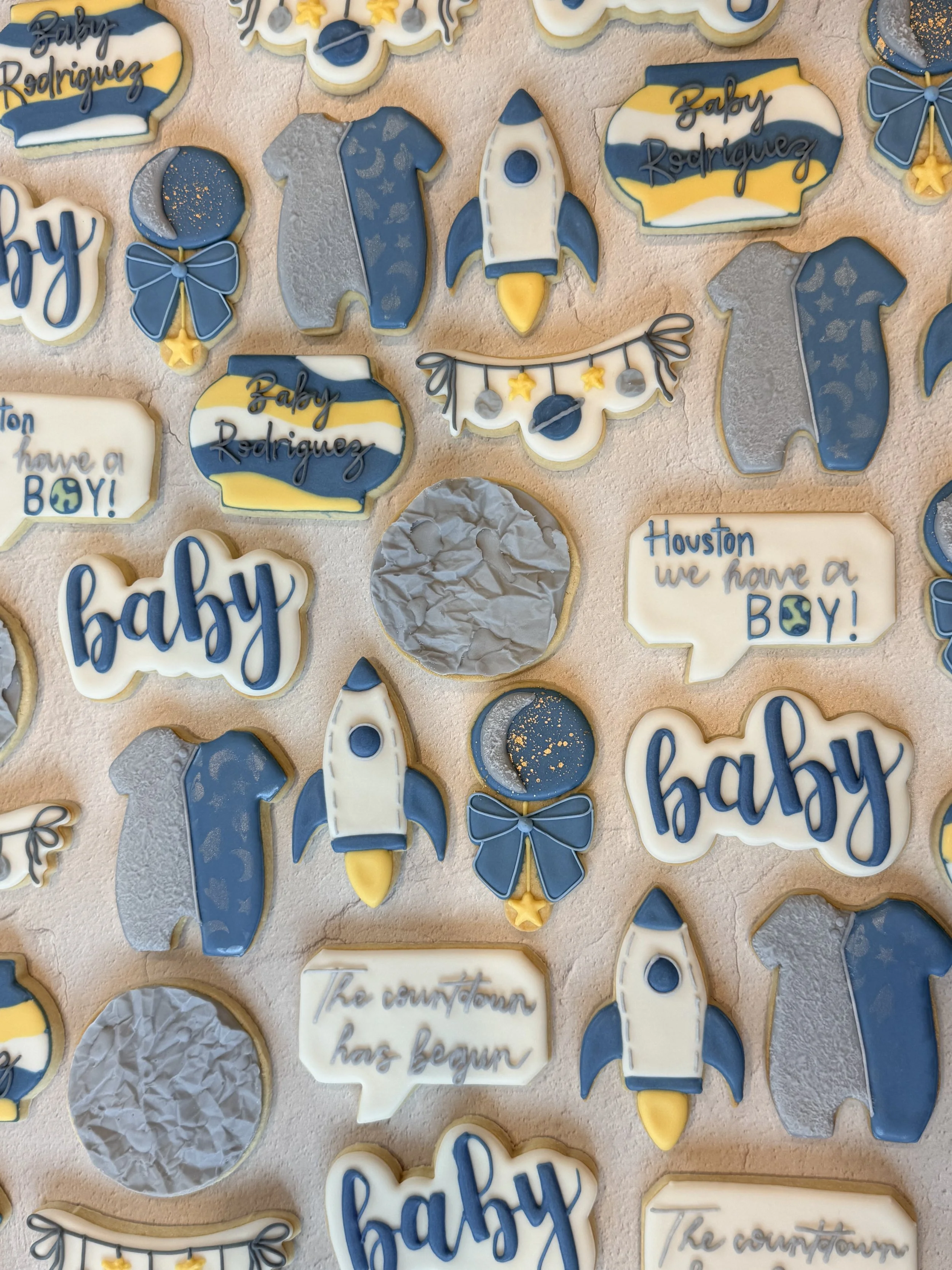 Collection of decorated cookies with space-themed designs, including rockets, planets, astronauts, and Newborn baby messages in blue, white, yellow, and gray colors.