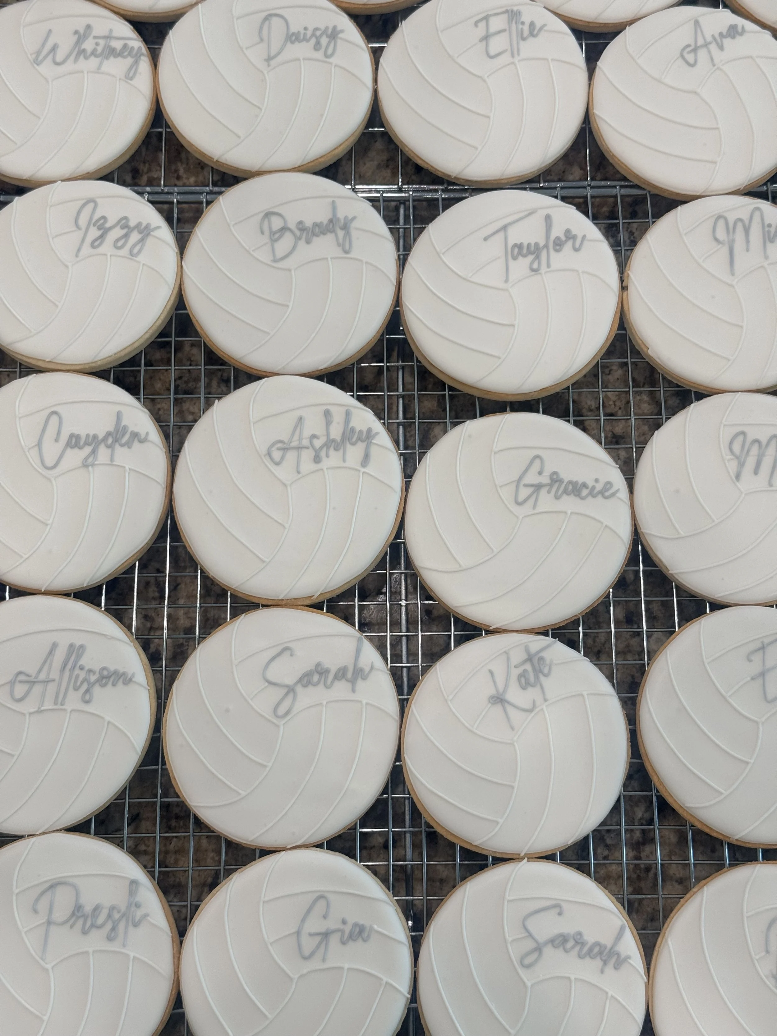 A collection of decorated cookies arranged on a grid, each with a white icing top and a handwritten name in gray icing, including names like Whitney, Daisy, Ellie, Avar, Izzy, Brady, Taylor, Alison, Sarah, Kate, and Gracie.