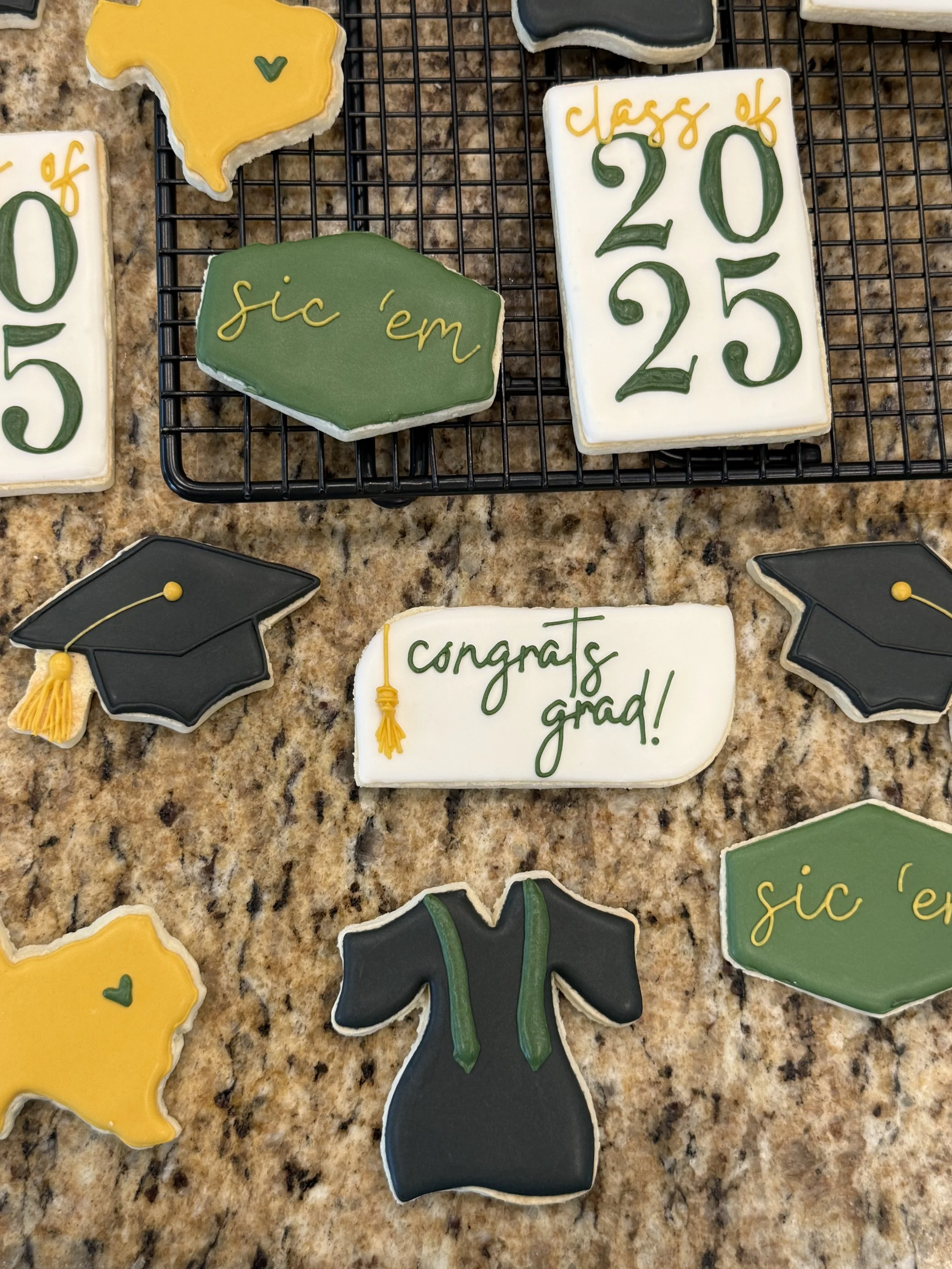 Decorative cookies on a countertop, including a green graduation gown, black cap, and white rectangular cookie with green and yellow writing that says "congrats grad!"
