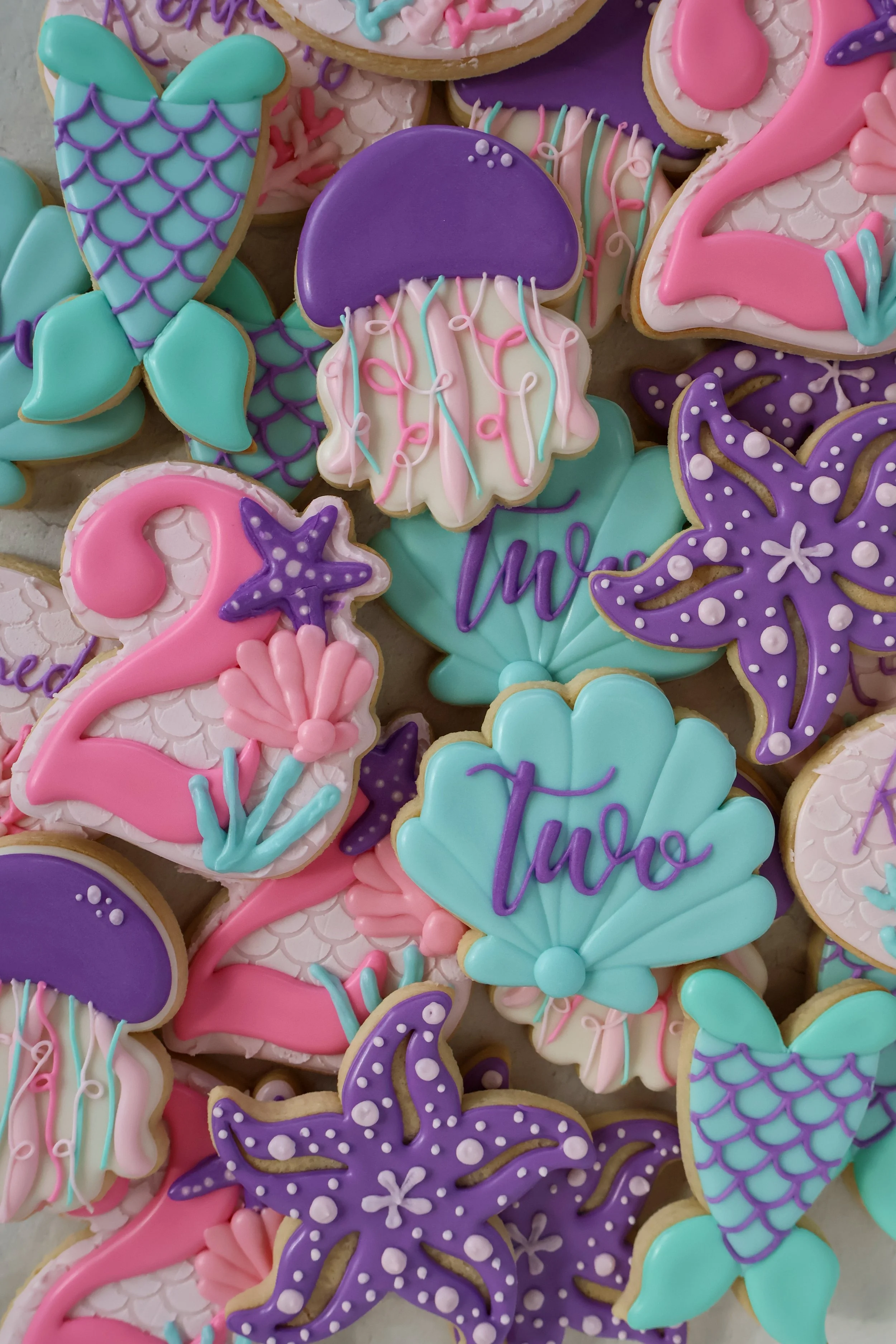 Colorful mermaid and starfish shaped decorated cookies with pink, purple, white, and turquoise icing, featuring sea-themed designs and text, arranged closely together.
