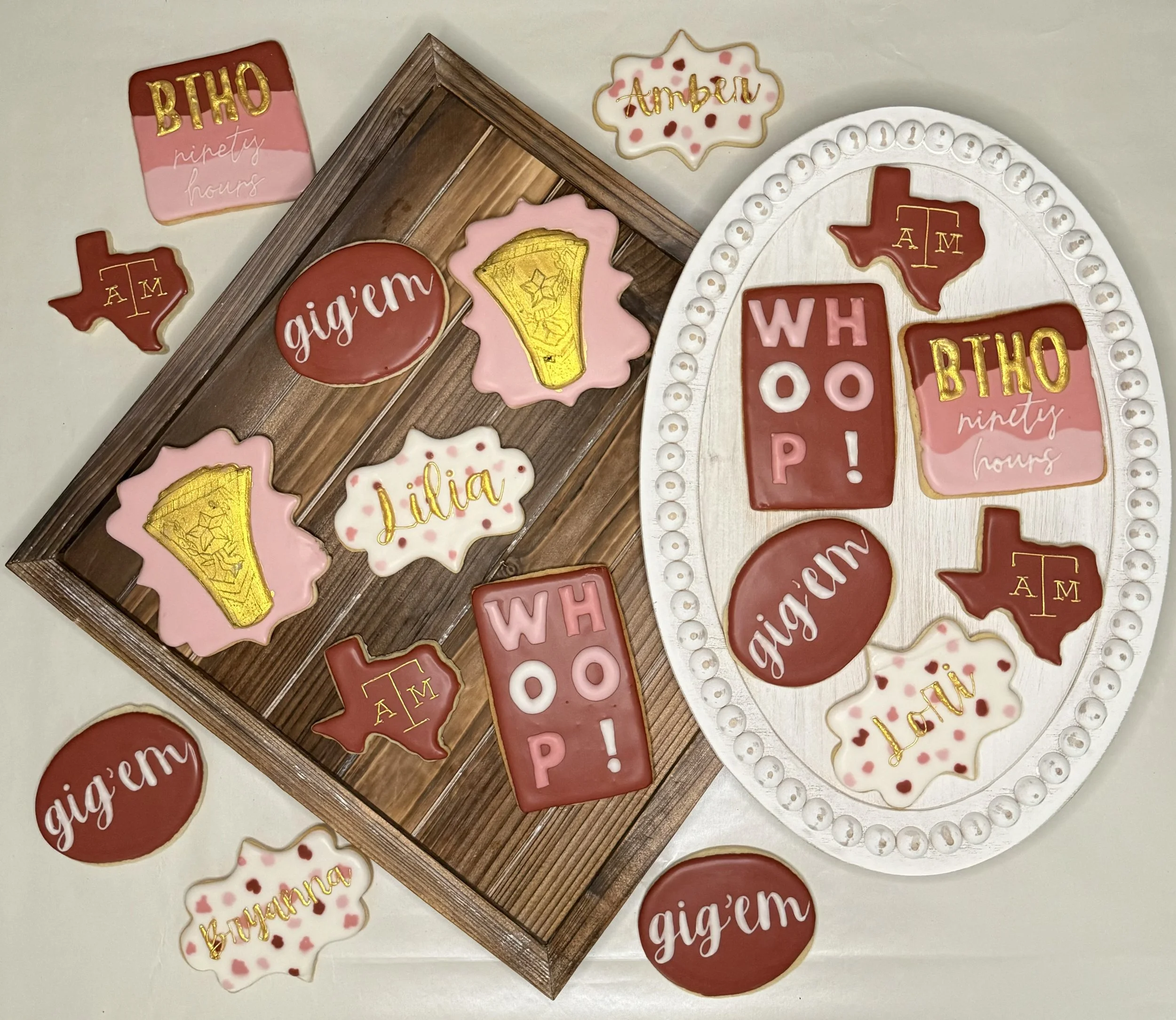 Decorative cookies with Texas-shaped cutouts, some with the text 'AM', 'WH', and 'BHO', others with floral patterns, gold accents, and names like 'Julia' and 'Amber', arranged on a wooden tray and a white decorative board.