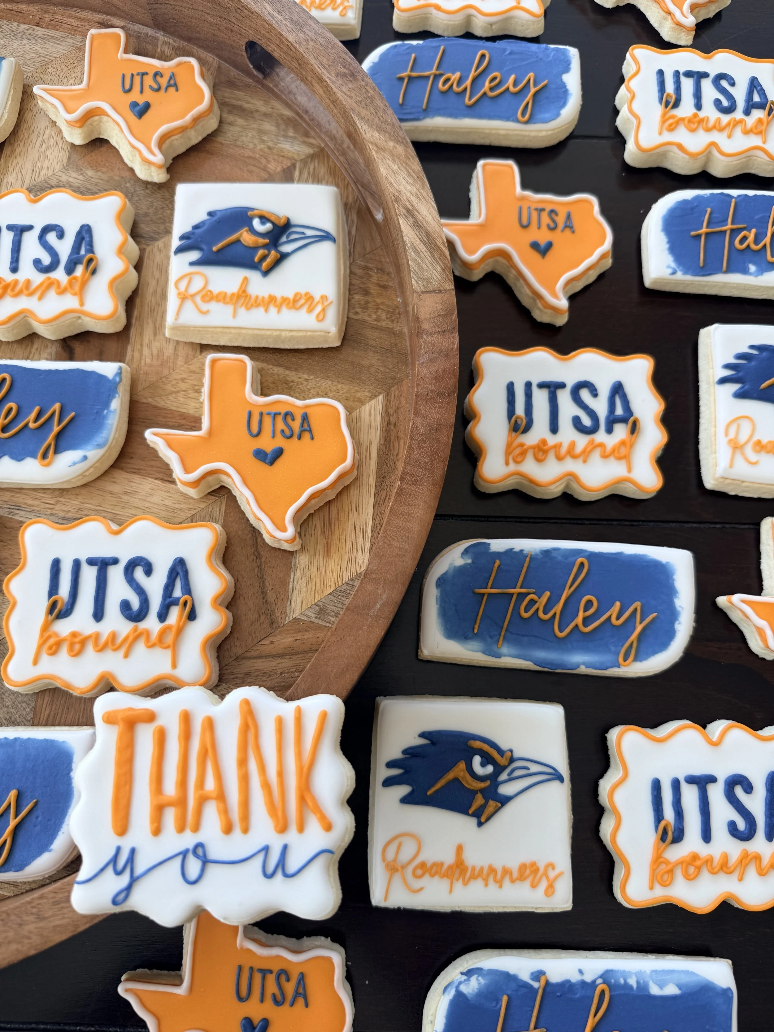 Decorative sugar cookies on a black surface and a wooden tray, featuring university of Texas at Austin-themed cookies with the words 'UTSA,' 'UTSA round,' and 'thank you,' along with cookies shaped like the state of Texas and a bird logo, all decorat