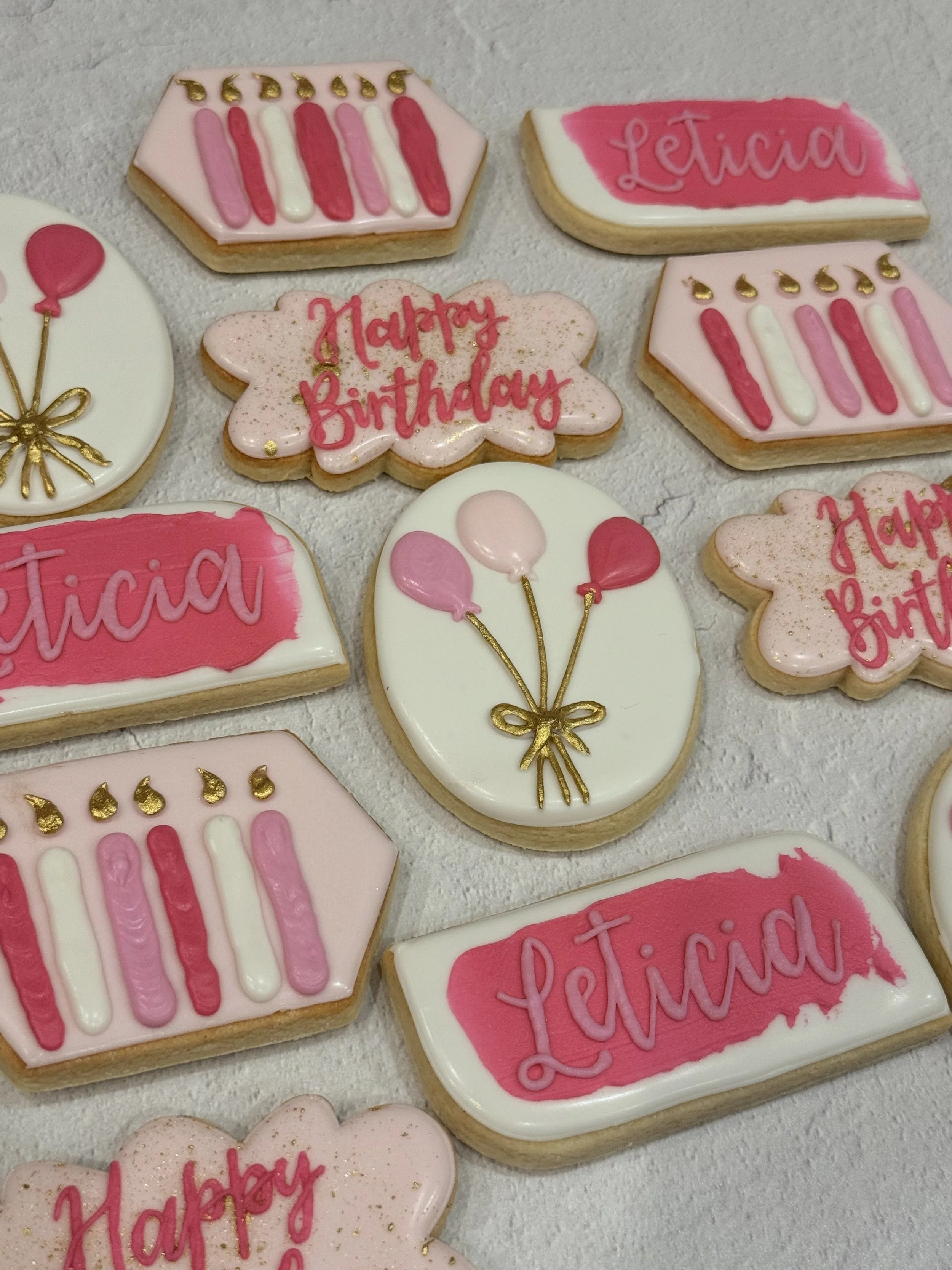 Assorted decorated birthday cookies with pink, white, and gold accents, featuring messages like "Happy Birthday" and "Leticia," and designs such as balloons and candles.