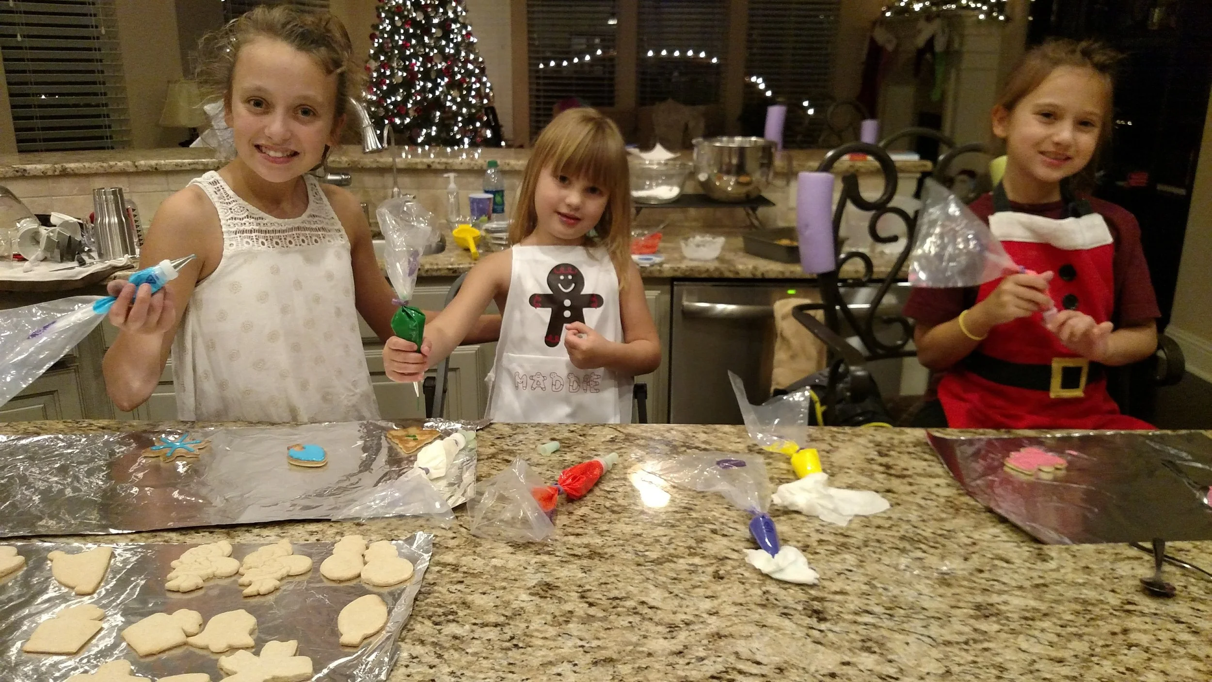 Three young girls decorating Christmas cookies in a kitchen with a Christmas tree in the background. They have icing and cookies on the counter.