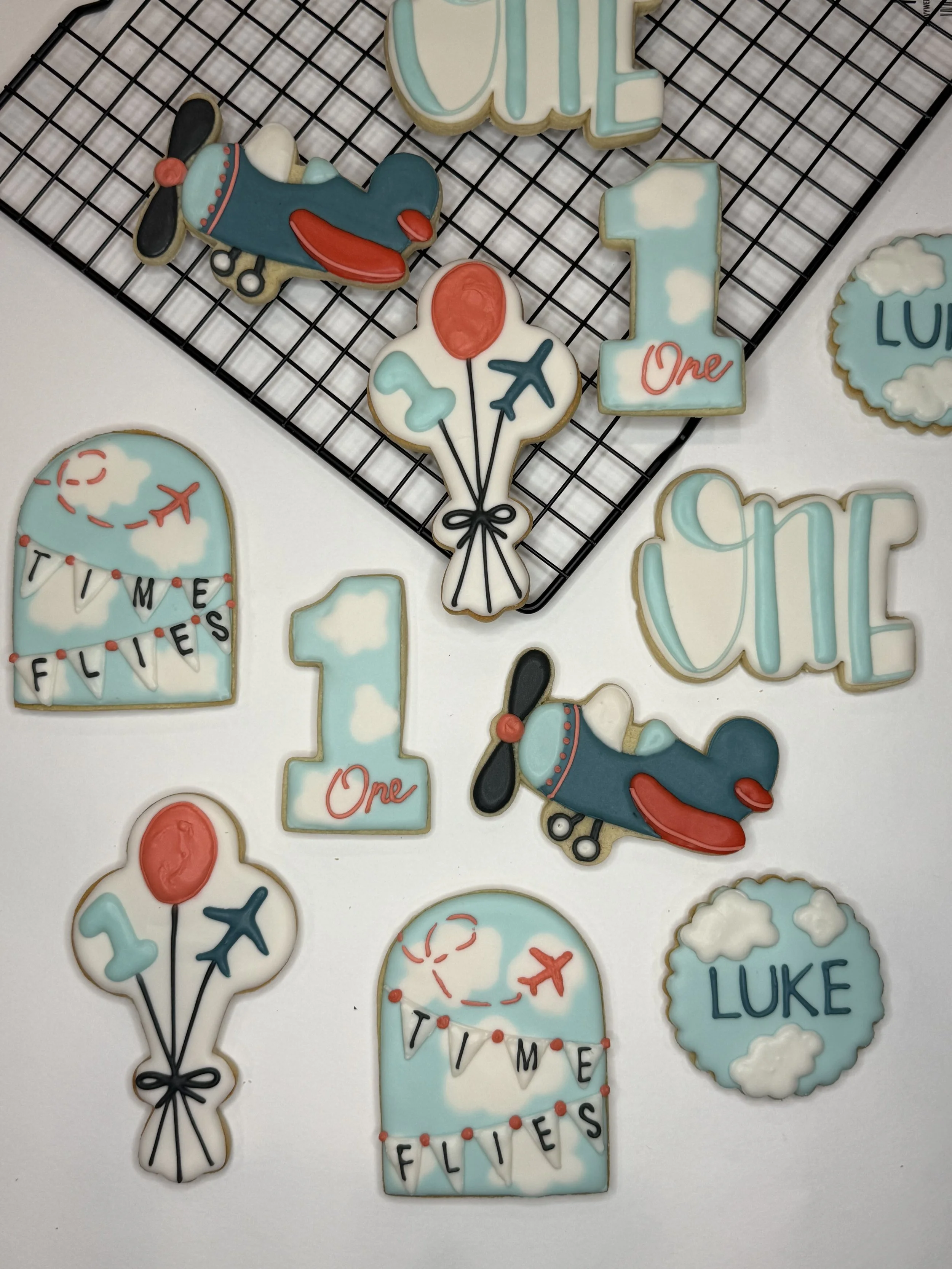 Cookies decorated with pastel blue, white, and red icing, including airplanes, balloons, the number one, and words like 'Time Flies' and 'Luki' for a first birthday celebration.