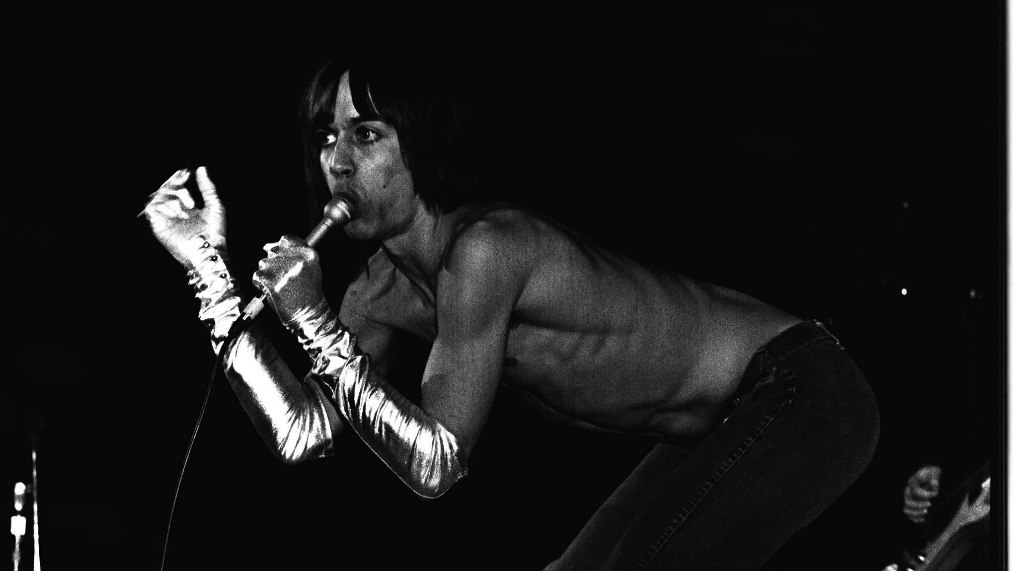 Black and white photo of a woman singing into a microphone, wearing shiny arm gloves, with her hand cupping the microphone. She is leaning forward on a dark stage.