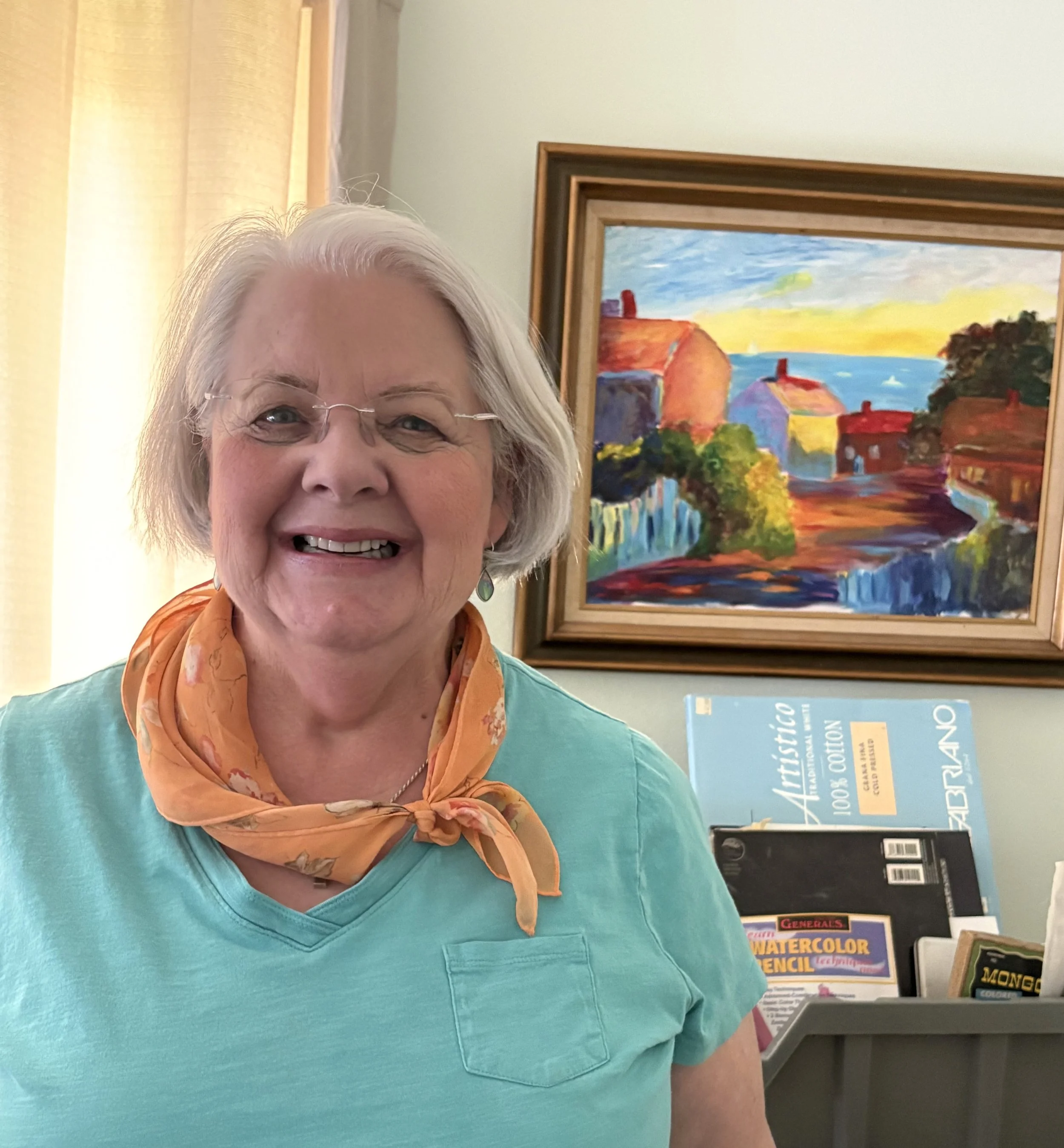 Close-up of smiling older woman with gray hair, wearing glasses, an orange scarf, and a turquoise top, standing in front of colorful artwork and books on a bookshelf.