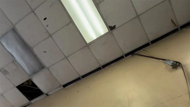 Ceiling tiles with some missing or damaged, fluorescent lights, and an electrical outlet with cables on a beige wall.