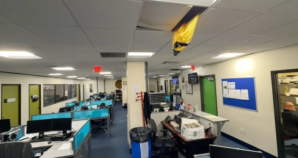 Office space with rows of workstations, some with cubicles, and several computer monitors. There is a partial ceiling collapse with yellow insulation hanging down. The room has a blue and white color scheme, with a green door and a bulletin board.