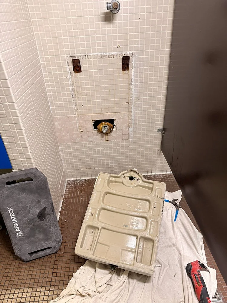 Bathroom stall with removed toilet, exposing plumbing; tools and equipment are on the floor, including a towel, a red power drill, and a black toolbox.