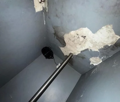 A damaged ceiling with peeling paint and exposed material in a corner of a room, with a metal pole and a black electrical box visible.