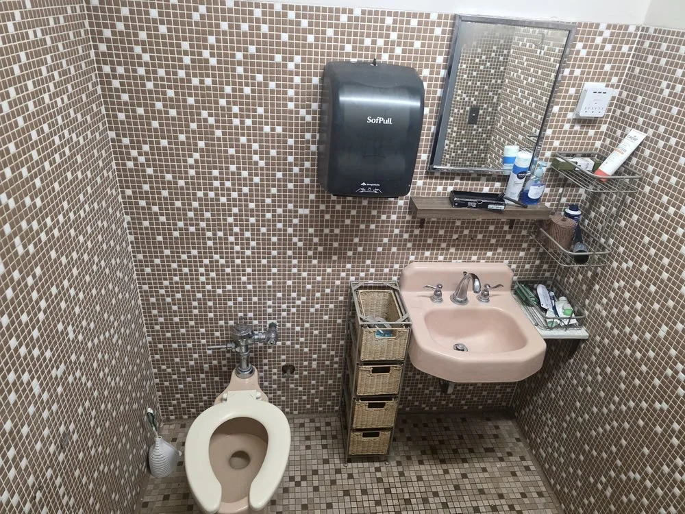 Small bathroom with mosaic tile walls and floor, pink sink, mirror, soap dispenser, and shelves with toiletries, including a black soap dispenser, a small wicker basket, and cleaning supplies; a paper towel dispenser on the wall; a toilet brush besid
