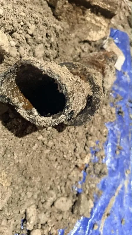 Close-up of a corroded metal pipe lying on the ground with dirt and a blue tarp nearby.