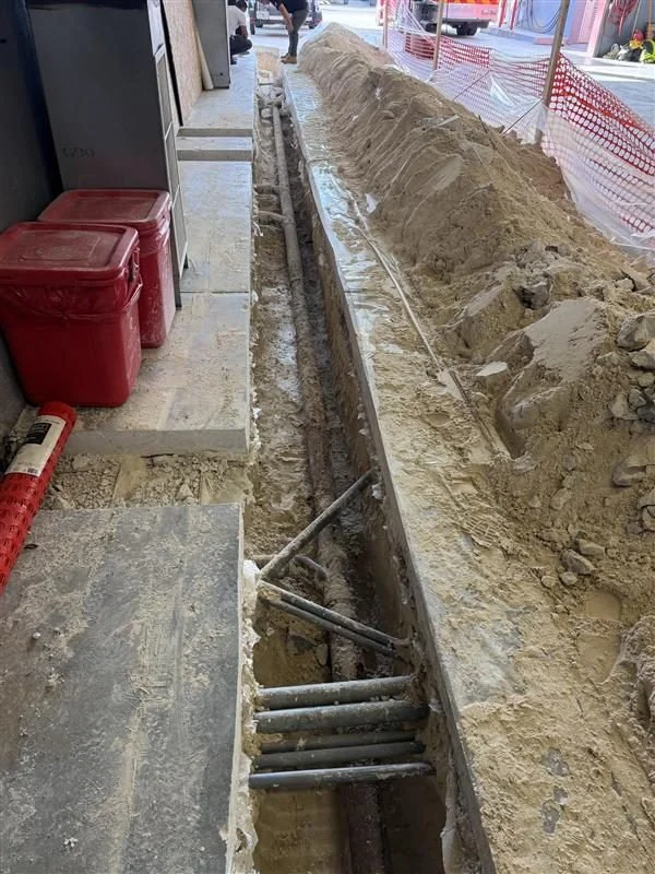 Construction site with a long trench filled with pipes, sand piled beside the trench, surrounded by orange safety fencing.