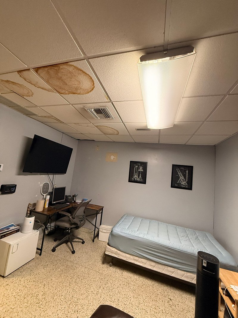 A room with stained ceiling tiles, a bed with a light blue mattress, a desk with a chair, a wall-mounted TV, and a trash can. The ceiling has visible water stains.