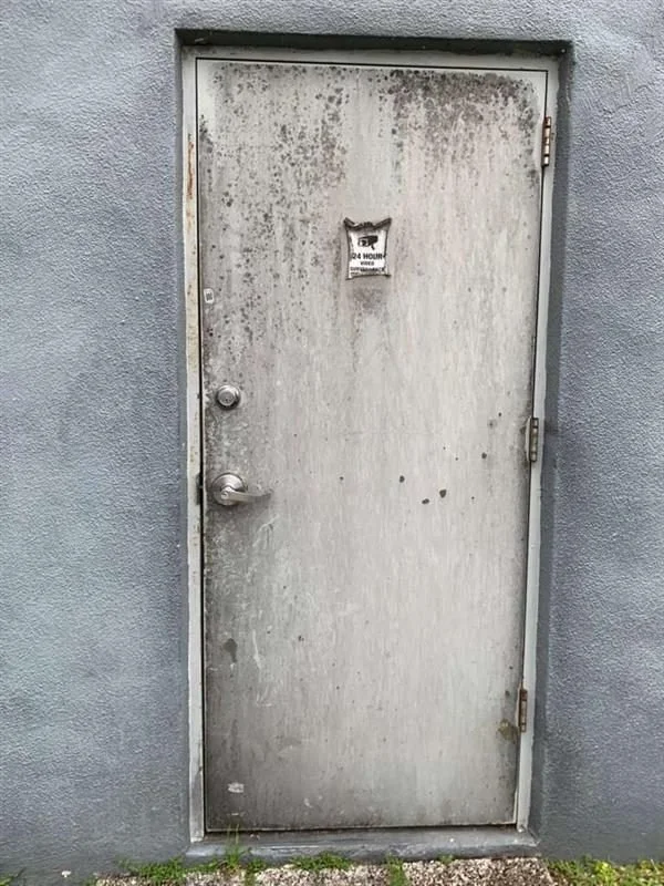 A weathered gray metal door with a small posted flyer, set in a blue wall. The door has a round doorknob and hinge on the right side, with some rust and dirt stains.
