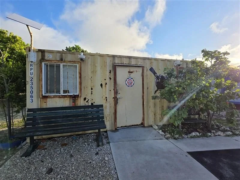 A rusty, small, tan-colored building with a door, window with metal bars, and security cameras outside. There is a bench in front and some greenery with trees nearby. The building has a sign with a logo and a mural of a large black gun on the right s