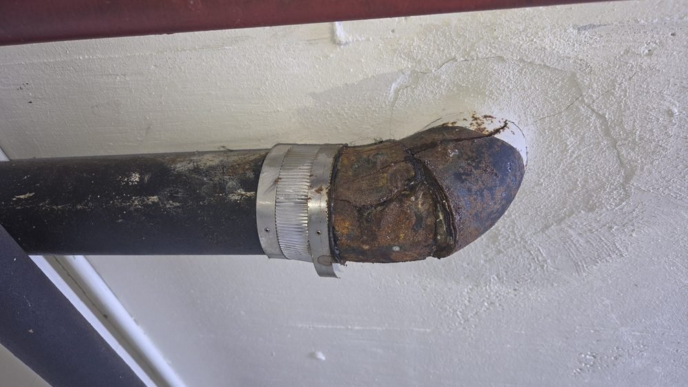 A corroded pipe with a large rusted section ending in a broken pipe tip, attached to a wall with a metal clamp.