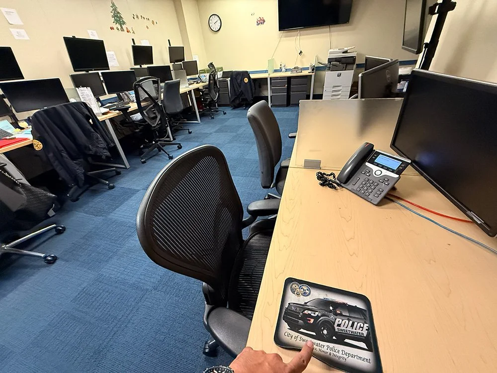 An office with multiple computer desks and chairs, a large flat-screen monitor, a desk phone, and a police department badge on the desk in the foreground.