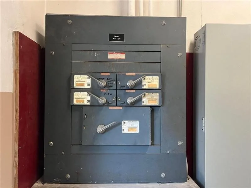 An electrical distribution panel with multiple switches and labels, mounted on a wall with red and beige paint.
