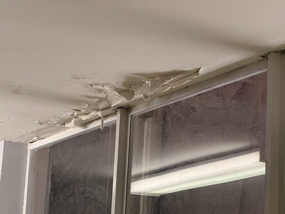 Ceiling paint peeling near a window, revealing damaged drywall.
