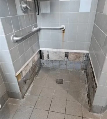 A partially renovated shower stall with exposed lower wall tiles, a drain on the floor, and damaged wall tiles near the floor. There are safety grab bars on the wall.