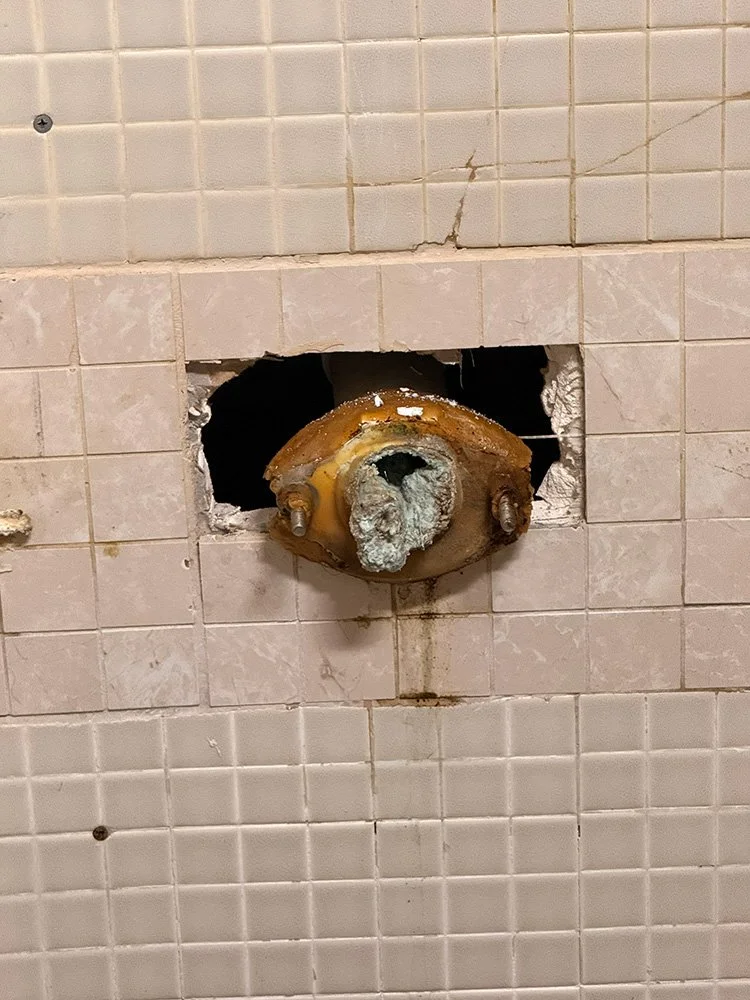 Wall with damaged tiles and an exposed rusted pipe with mold or mineral buildup.
