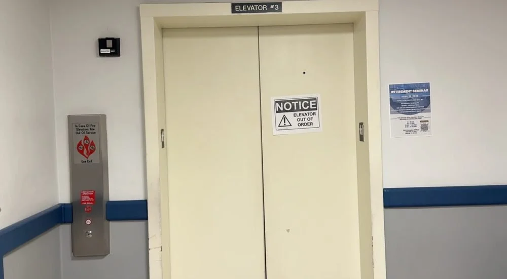 Elevator door with a sign reading "NOTICE Elevator Out of Order" in a hospital or public building corridor, with a fire alarm panel on the left and a poster on the right.