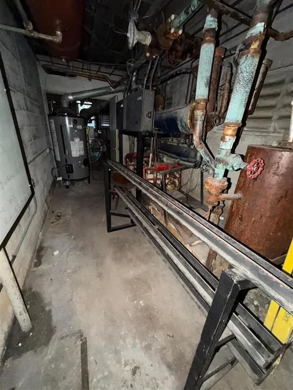 An industrial utility room with rusted and corroded pipes, a large tank, and electrical control panels. A metal track or rail runs along the floor.