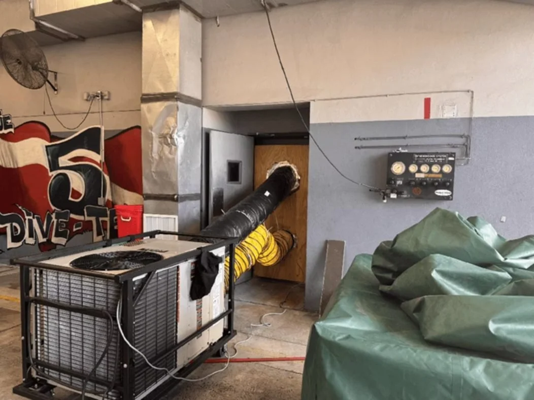 An indoor scene with HVAC or industrial ductwork connected to a wooden board through a large black flexible pipe, a large green tarp covering objects on the right, and a red and black mural with the text 'DIVE', suggesting a dive shop or aquarium the