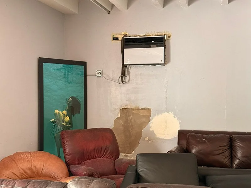 A room with three leather armchairs and a leather sofa, a large painting of a person underwater holding a yellow flower, and an air conditioning window unit on the wall with exposed wiring and unfinished wall repair.