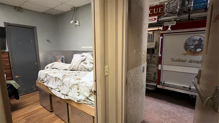 An unmade bed with white sheets and blankets in a small room, partially visible, with a door and some furniture nearby. Outside, a fire truck is parked outside, visible through an open doorway.