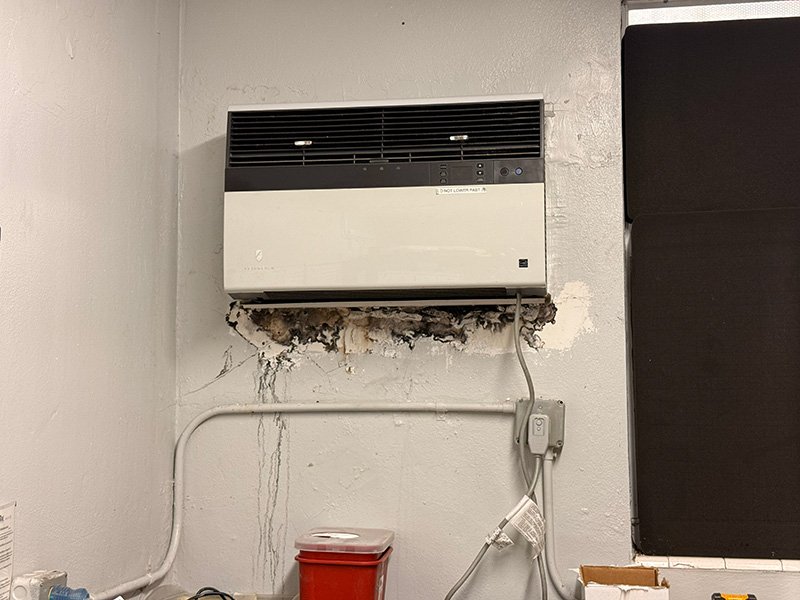 An air conditioning unit mounted on a wall with visible damage and mold beneath it, a power outlet and a red container below.
