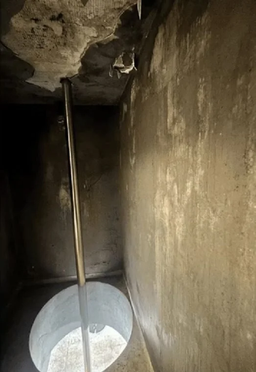 A pole inserted into a hole in the ground inside a small, dark, concrete-walled room with a rough, damaged ceiling.