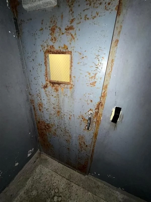 Rusted metal door with a small window, handle, and an electronic lock, in a dark, worn hallway corner.