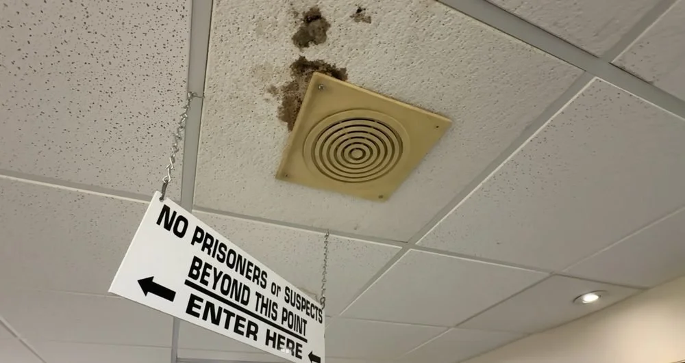 Ceiling with a yellow vent and a sign that says, "No prisoners or suspects beyond this point. Enter here" with an arrow pointing left.