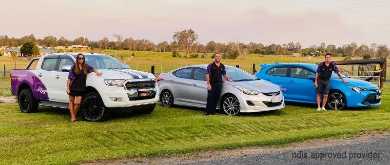 Three people standing next to three different cars parked on a grassy field in a rural area.