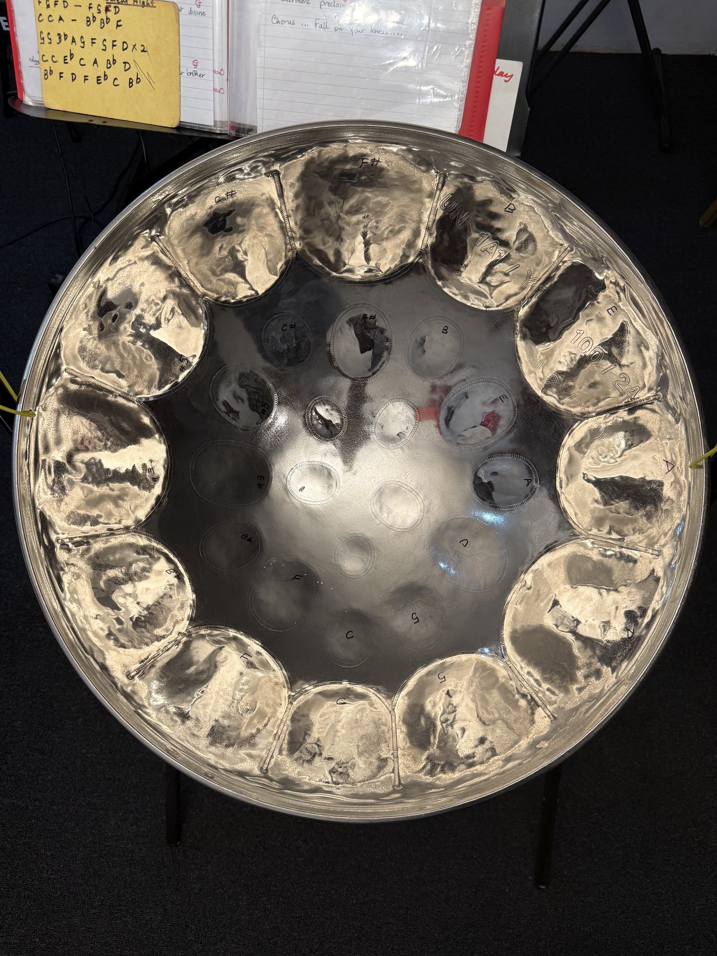 A large metallic handpan drum with labeled notes. Handpan has several circular indentations, each marked with musical notes and letters.